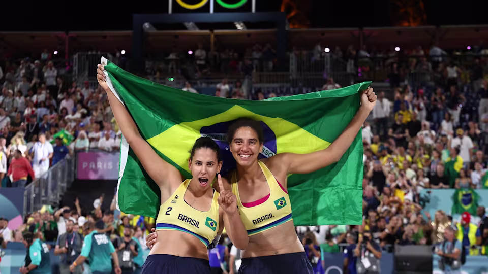  A dupla brasileira feminina de volei de paria juntas segurando a bandeira do Brasil em comemoração da conquista do ouro em Paris logo apos o set