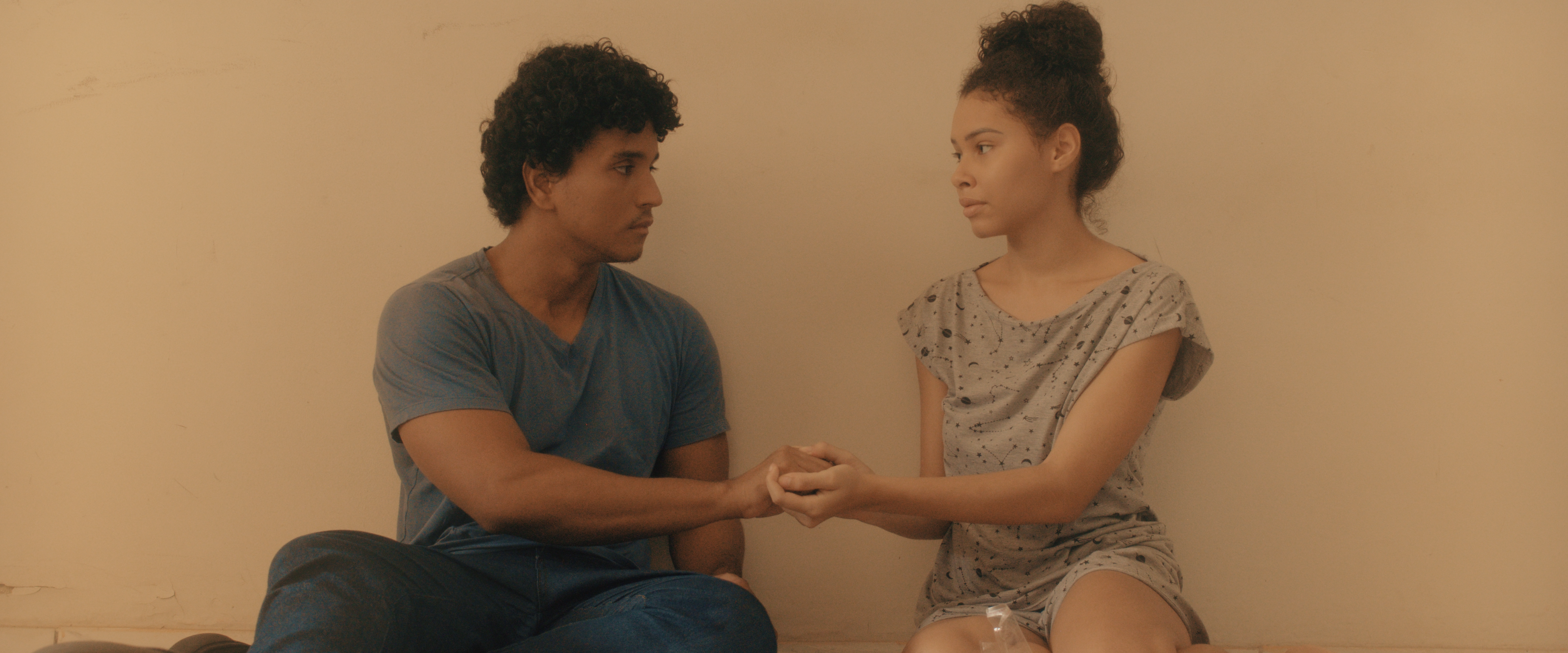 Julia e João, personagens irmãos do curta sentando na sala vazia, olhando um para o outro