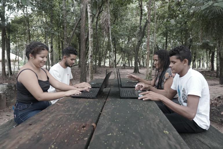 jovens sentados à mesa com computadores no meio da floresta