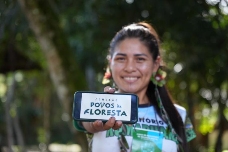 Indígena mostrando o celular com a inscrição "conexão povos da floresta"