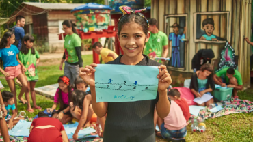 Garota sorrindo segurando desenho, ao fundo outras crianças fazendo atividades recreativas