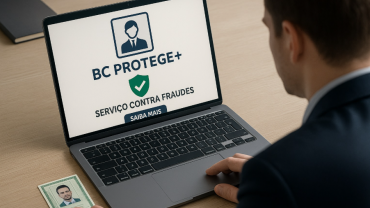 Homem em frente a um notebook ligado sobre Uma mesa. Na tela do PC há uma imagem onde se lê "BC PROTEGE + SERVIÇO CONTRA FRAUDES".