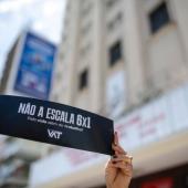 Paisagem urbana, em foco: levantada por mãos, uma faixa preta com a mensagem "Não a escala 6x1".