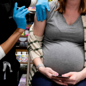 A imagem mosta uma mulher grávida sentada recendo uma dose de vacina no braço direito.